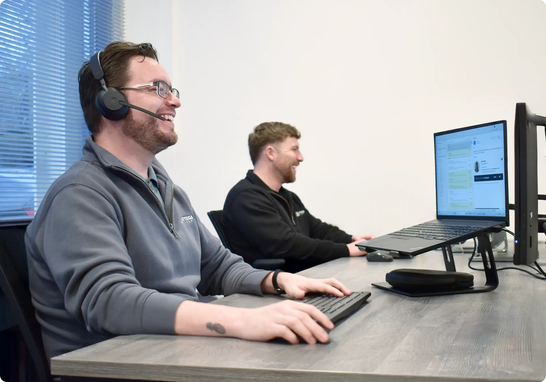two men from the attega team smiling whilst looking at their computers, both sat on the same desk